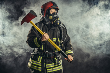 Obraz premium portrait of serious and confident caucasian fireman stand holding hammer, wearing special protective uniform in the smoky background