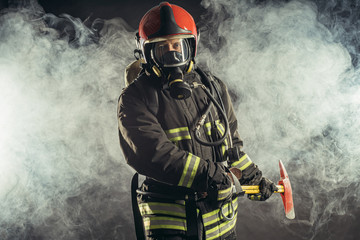 Obraz premium portrait of serious and confident caucasian fireman stand holding hammer, wearing special protective uniform in the smoky background