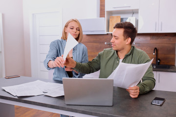 young married couple irritated while sitting on table at home, using laptop and holding reading documents