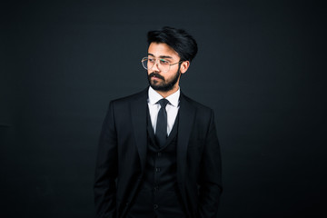 Portrait of a young handsome successful Indian in a classic black three-piece business suit on a dark background, hold down the glasses