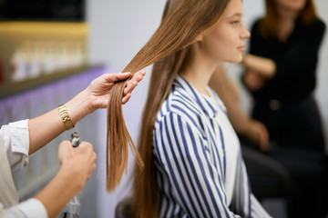 Obraz premium side view on beautiful caucasian girl sitting on chair in beauty salon, closeup of long hair, hairdresser hands holding them, going to cut split ends. Beauty, hair concept