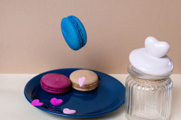 Obraz na płótnie Canvas Sweet blue macaroon falls into a plate on a light beige background with a glass can and a heart-shaped lid. Dessert.