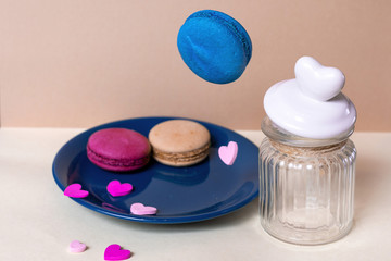 Obraz na płótnie Canvas Sweet blue macaroon falls into a plate on a light beige background with a glass can and a heart-shaped lid. Dessert.