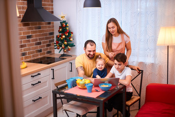 adorable kids and careful parents sit in kitchen with laptop, watching films and videos, eating fruits, having breakfast together at morning