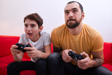 virtual, video game. caucasian bearded man playing video games with son indoors, sitting on sofa,...