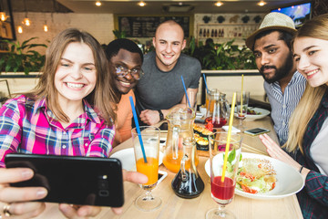Group of young cheerful friends are sitting in a cafe, eating, drinking drinks. Friends take selfies and take pictures.