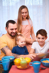 Fototapeta na wymiar lovely adorable happy caucasian family consisted of young mother with long hair, bearded father and little kids son and daughter sit together on table, using laptop and eating, having meal, breakfast