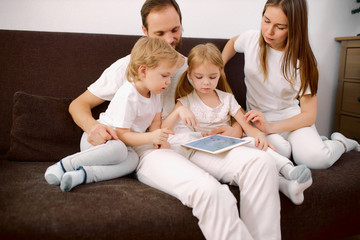 young caucasian family watch video on modern tablet sitting on sofa together, dressed in white casual clothes, look at screen of tablet