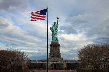 Statue of Liberty from behind with waving flag of the United States of America in autumn and cloudy weather