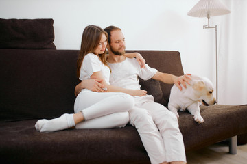 portrait of adorable caucasian couple sit on sofa with friendly white dog, people and animals concept. young man and woman in white casual clothes