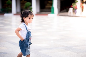 Side view of cute Asian little girl walking around the courtyard of Wat Sutad. A 2 years old in a blue denim overalls. Concept of taking children on a trip.