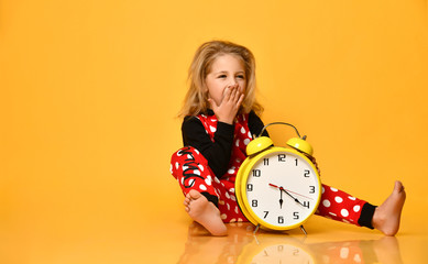 Little blond girl red dotted home clothing sitting on floor with big yellow alarm clock and yawning over yellow background