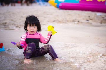 An Asian girl was sitting in the sand and holding a sand toy in her hand. A cute kid is wearing a purple-black bathing suit on the beach. Children 2 years old. © Kanthita
