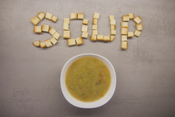 word SOUP made of square toast ryados with a round bowl of soup on a gray kitchen table with texture top view