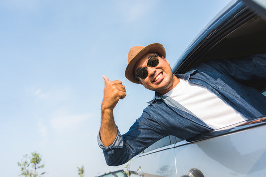A traveler wearing a hat is driving a car and then pulling himself out of the car window. And thumbs up This picture is about a safe journey by car.