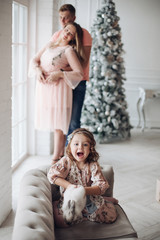 Stock photo of lovely girl playing with bunny on the sofa. Her parents standing snuggling in the background next to decorated Christmas tree.
