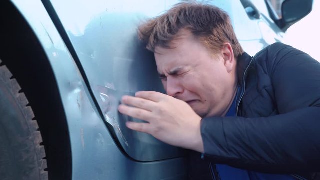 Broken automobile in parking lot. Guy is crying hysterically because of dented car door, beating it with his fist. Expression of emotions of sadness, despair, hopelessness and grief, the close-up.