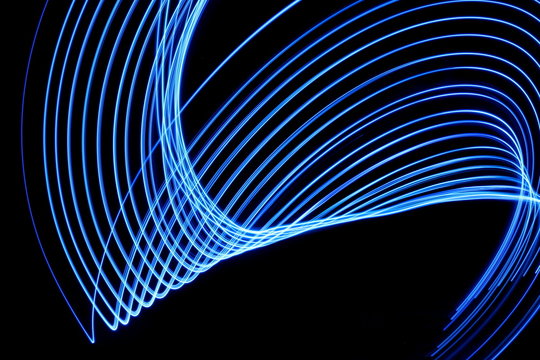 Long exposure photograph of neon blue colour in an abstract swirl, parallel lines pattern against a black background. Light painting photography.