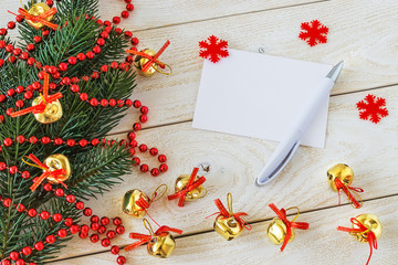 Empty greeteing card and ballpoint pen on a wooden table near a fir branch with red bead garland and golden Christmas balls. Christmas and New Year holydays.