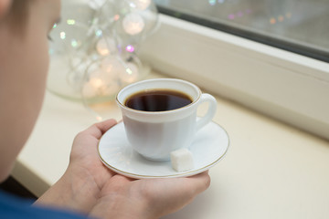 the child stands near the windowsill and holds a cup of coffee next to the lights
