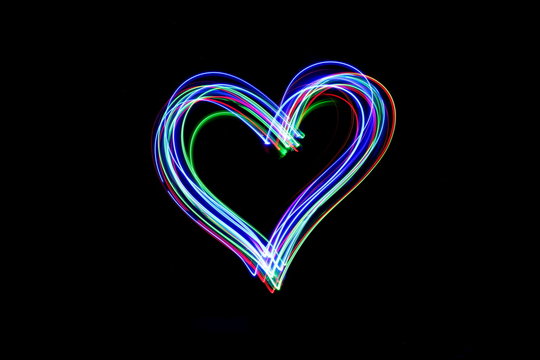 Long exposure photograph of a heart outline in neon colour in an abstract swirl, parallel lines pattern against a black background. Light painting photography.