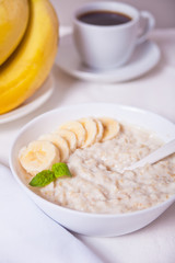 Oatmeal porridge with banana in the white bowl, cup of coffee on the white table. Health breakfast.