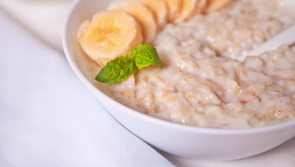 Oatmeal porridge with banana in the white bowl on the white table. Health breakfast. Close up.