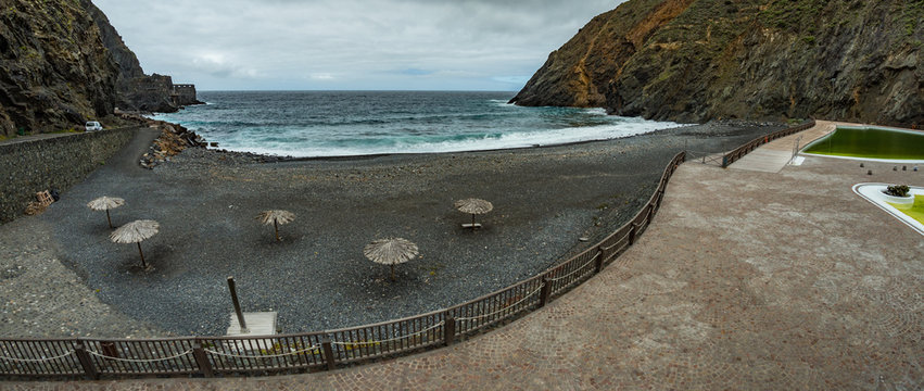 Wide panorama. Volcanic black sand. Beach umbrellas and a children's pool. Vacation spot for locals and tourists. Castillo del Mar - old banana factory in the background. Vallehermoso, La Gomera