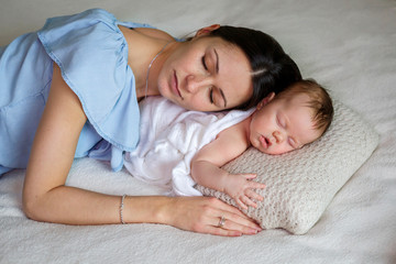 Young mother holding her sleeping newborn baby in her arms in light bedroom. Family, motherhood, love, health and care