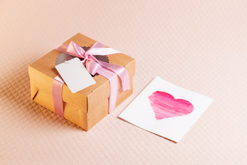 A square gift box with a pink ribbon on a light pink background and label for space for text. A card with a heart that is painted with watercolors