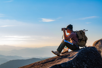 A young man with a lifted backpack looked in binoculars sitting on top of the mountain enjoying a spectacular view on the mountains above the clouds. - Powered by Adobe