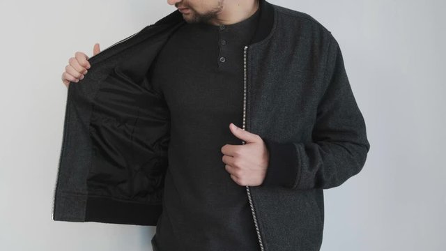 A young man in dark clothes zips up his jacket standing near a white wall.