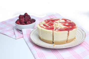 cut cheesecake on a plate and a bowl of raspberry on red and white tablecloth isolated on white background