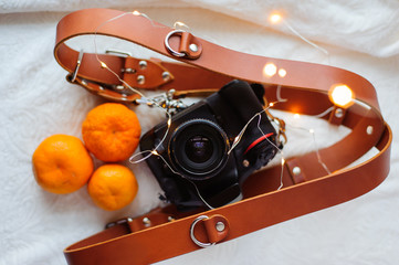 Photo camera, leather straps, tangerines and christmas lights on the white background.