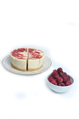 cut cheesecake on a plate and a bowl of raspberry isolated on white background