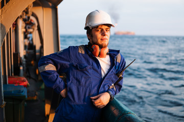 Marine Deck Officer or Chief mate on deck of offshore vessel or ship , wearing PPE personal protective equipment - helmet, coverall. He has VHF walkie-talkie radio