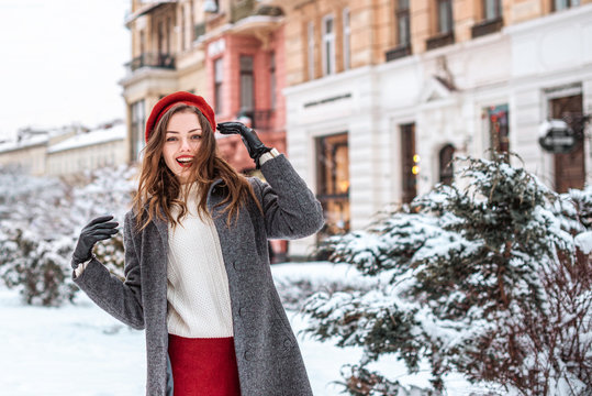 Happy smiling girl enjoying winter holidays, walking in snow covered street of European city. Model wearing red beret, white sweater, grey coat. Copy, empty space for text