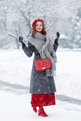 Happy smiling girl enjoying snowfall, winter holidays, walking in snow cowered street of European city. Model wearing red beret, skirt, boots, grey scarf, coat, with quilted bag