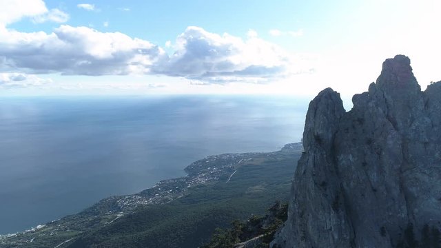 Aerial view of sharp rocky cliffs above blue ocean on cloudy sky background. Shot. Flying over the mountain peaks and coastal small city.
