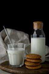Transparent glass of milk, a bottle of milk and oat cookies on brown cutting board