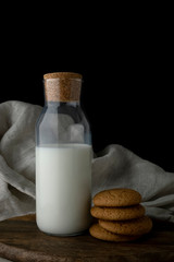 Transparent bottle of milk and oat cookies on brown cutting board, textile background