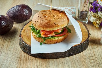 Tasty burger with salmon, tomatoes, arugula and cucumbers on a wooden board on a wooden background. Fishburger. Healthy snack. Close up view