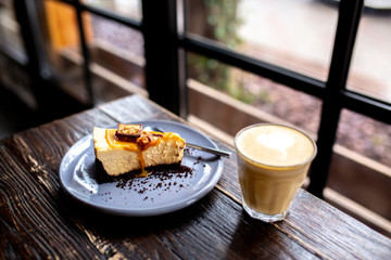  Slice of cheesecake with a cup of coffee on a wooden table. Cheesecake with nuts and caramel. Snickers cheesecake. Caramel Pie. Slice  caramel cheesecake with popcorn. Dessert with latte in a glass