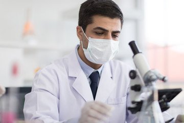 A male scientist wearing a mask and looking through the microscope in the laboratory