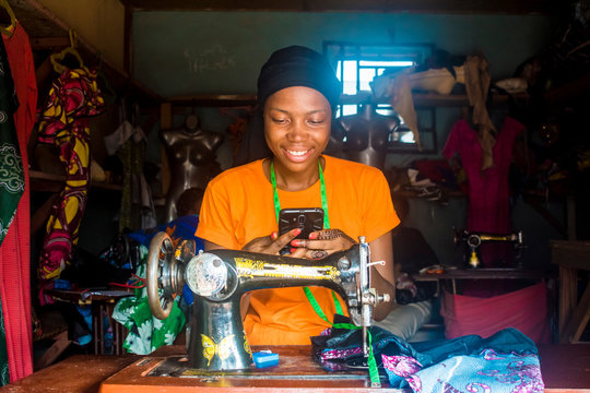 young african female tailor smiling while viewing content on her mobile phone sitting with her sewing machine