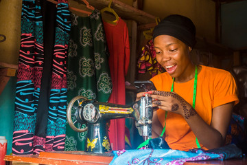 young african female tailor smiling while viewing content on her mobile phone sitting with her sewing machine
