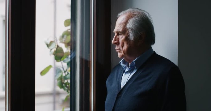 An elderly thoughtful smiling man is looking out a window of his house in the morning.