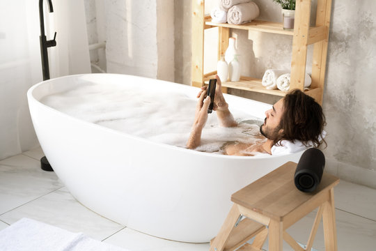 Young restful man scrolling in smartphone while taking bath with foam