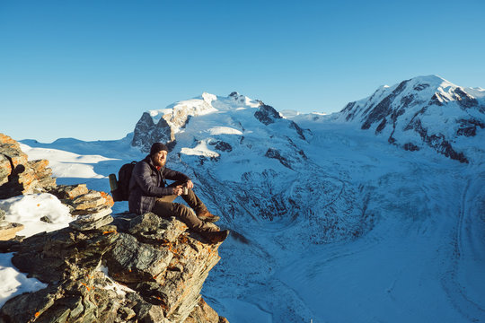Traveler Man with backpack trekking in mountains, enjoy beautiful Matterhorn view. Explorer man hiking on hills, travel in Swiss Alps, Switzerland. Hiker sitting on rock cliff outdoors on nature.