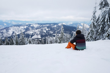 Traveler man sitting on the mountain hillside and looking on skyline view in front of beautiful pine trees in winter. Travel background with snow. Skier, explorer active holidays, vacation.